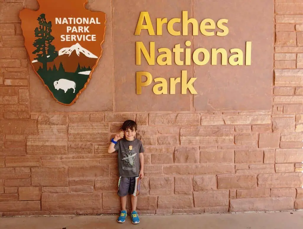 Eli in front of the Arches National Park Sign holding up his Junior Ranger Badge