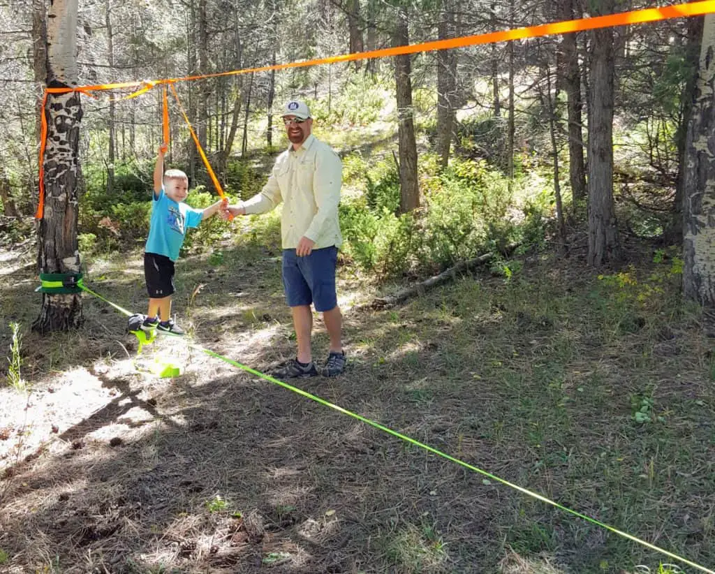 little boy walking on a slack-line in the woods next to his father