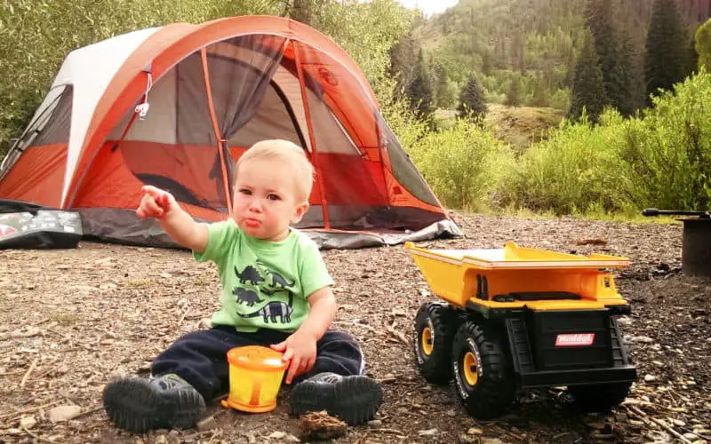 baby sitting in the dirt in front of a tent eating snacks