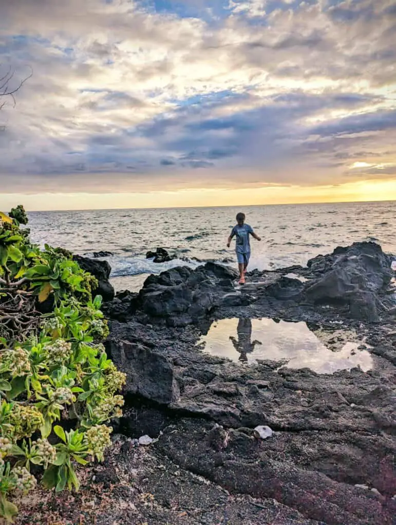 Boy standing on the edge of the ocean at sunset with his reflection in a tide pool